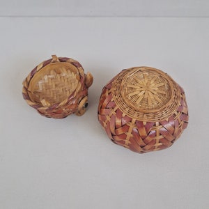 May include: A set of two woven baskets, one with a round shape and the other with a rounded, animal-like shape. The baskets are made of natural brown and red woven material.