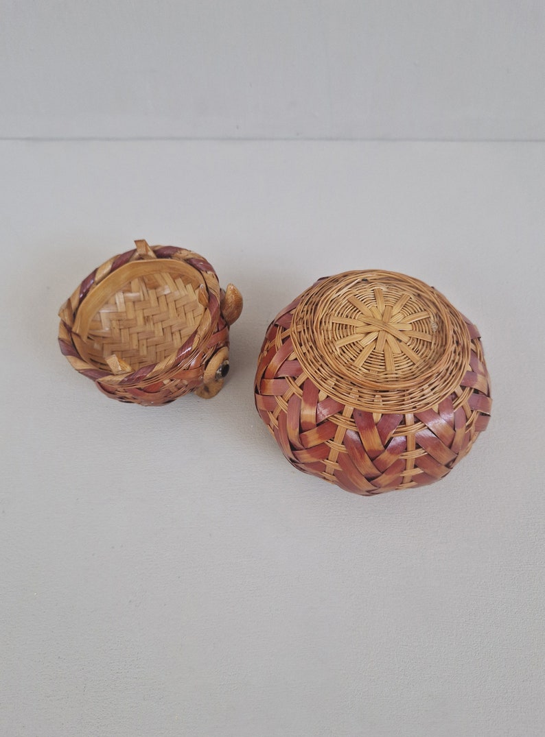 May include: A set of two woven baskets, one with a round shape and the other with a rounded, animal-like shape. The baskets are made of natural brown and red woven material.