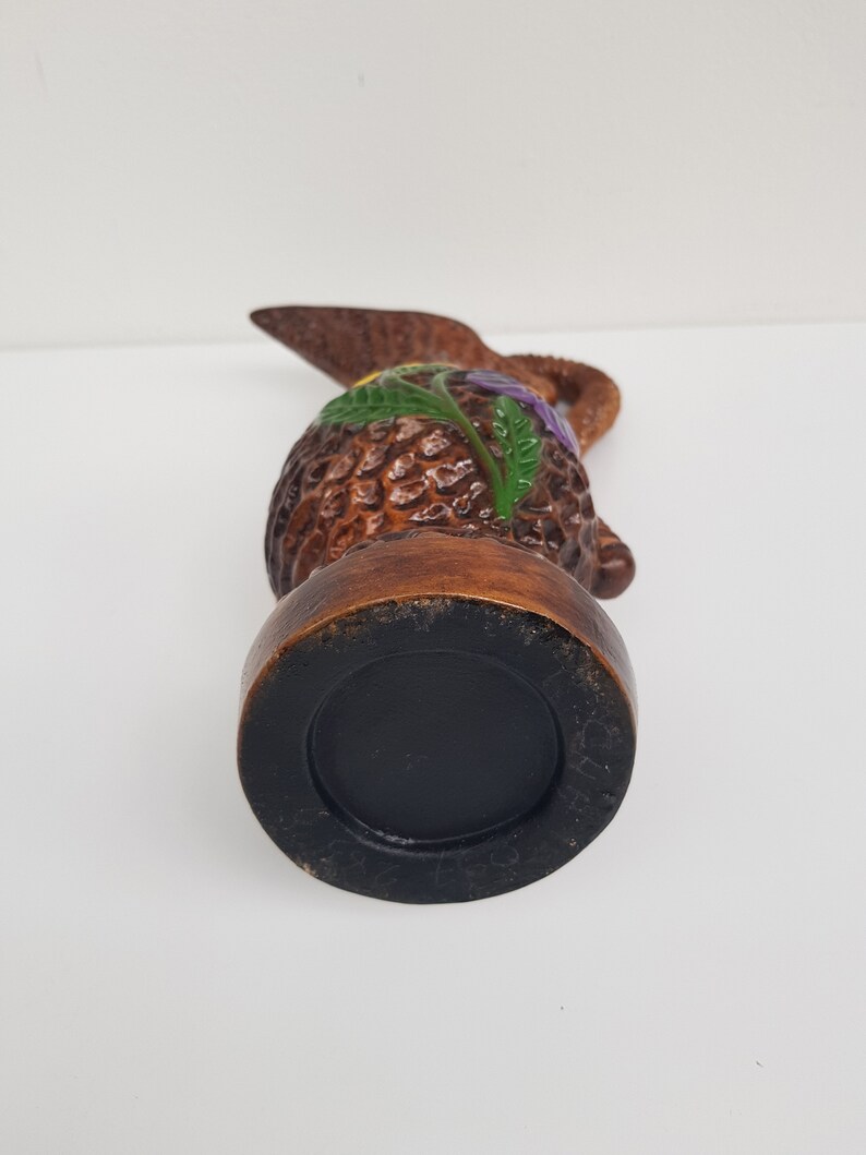 May include: A brown ceramic pitcher with a textured body and a handle. The pitcher is decorated with green, yellow, and purple floral accents. The base is black.