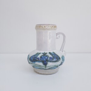 K&ouml;nnte beinhalten: Eine wei&szlig;e Keramikvase mit einem blauen und gr&uuml;nen Blumendesign. Die Vase hat einen Henkel und einen strukturierten Rand.