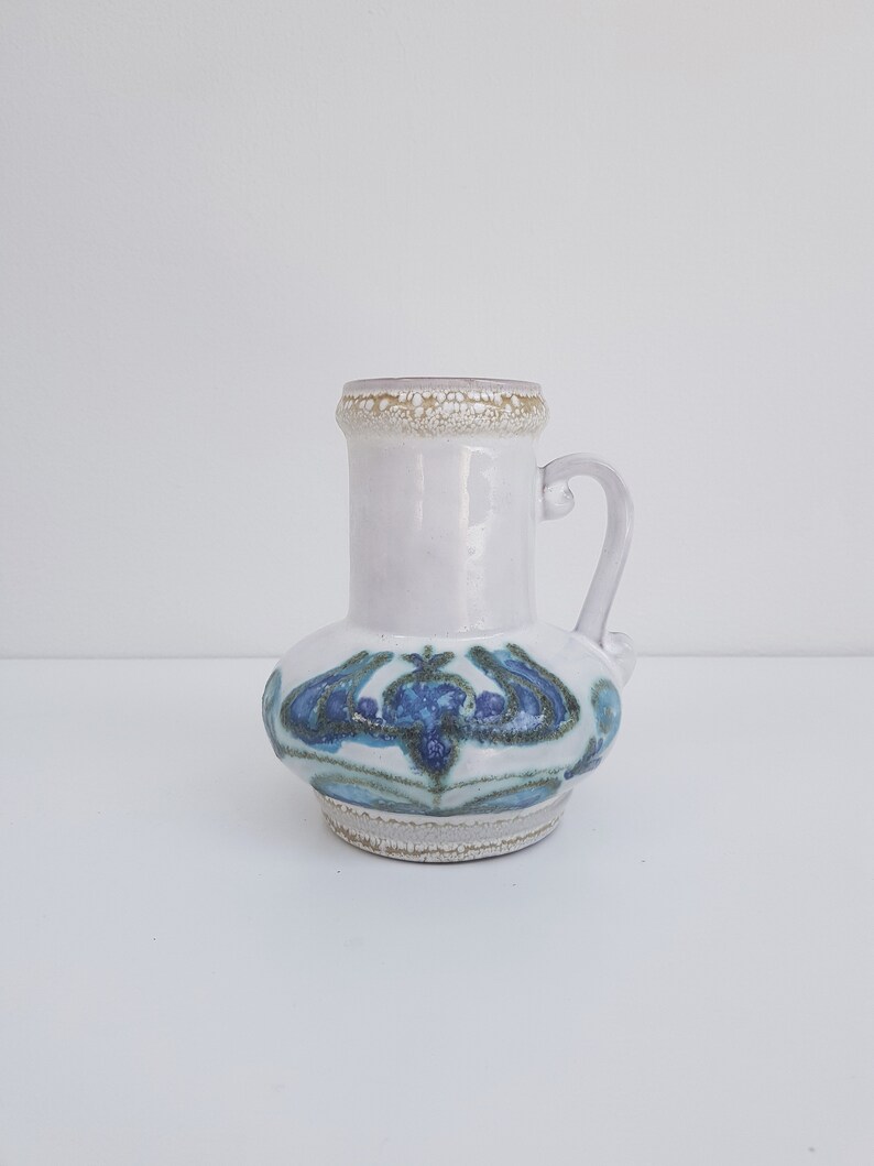 K&ouml;nnte beinhalten: Eine wei&szlig;e Keramikvase mit einem blauen und gr&uuml;nen Blumendesign. Die Vase hat einen Henkel und einen strukturierten Rand.