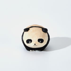 May include: A hand-carved wooden panda figurine. The panda has a light tan face and body with black accents around the eyes, ears, and body. The panda's eyes are black, and it has a small, carved mouth. The figurine is on a white background.