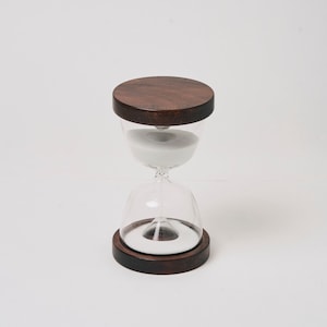 May include: A wooden hourglass with a clear glass bulb and white sand. The hourglass has a dark brown wooden base and top.