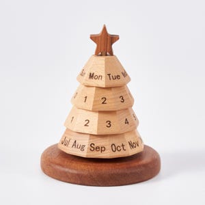May include: A wooden perpetual calendar shaped like a Christmas tree with a star topper. The light wood tiers feature black printed text for days of the week, numbers for dates, and months of the year, all resting on a dark circular wooden base.