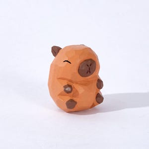 Puede incluir: Una pequeña figurita de madera tallada a mano de un capibara. La figurita es naranja con detalles marrones y tiene una cara sonriente.