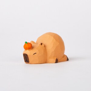 May include: A small, hand-carved wooden figurine of a capybara sleeping with a small orange pumpkin on its head. The figurine is painted in shades of brown and orange.