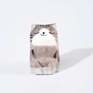 May include: A wooden cat ornament with a calm expression. The cat is mainly grey and white, with black stripes and markings. Its eyes are closed, and it has a small, smiling mouth. The ornament is carved from wood and has a simple, minimalist design.