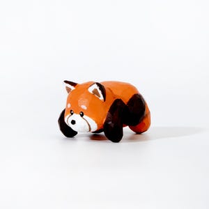 May include: A handcrafted wooden red panda figurine. The sculpture features a reddish-orange body with black legs and ears. The face is white with black eyes and a black nose. The figurine is posed in a lying down position.