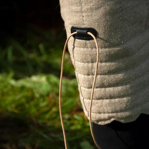 May include: A close-up of a beige quilted fabric with a black leather strap and a brown cord attached. The fabric is textured and has a slightly wavy edge.