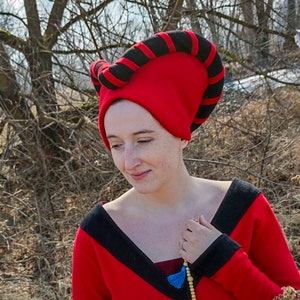 May include: A red medieval hat with black stripes and a matching red dress with black trim. The hat is a distinctive headpiece, likely for historical reenactment or costume use. The outfit is a vibrant combination of red and black.