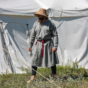 May include: A person wearing a grey tunic with a red belt and a straw hat stands in front of a white tent. The person is wearing brown trousers and brown shoes.