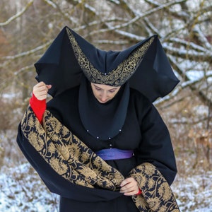 May include: A black headdress with a pointed, structured shape and gold floral embroidery, worn with a black garment featuring a wide, gold-patterned fabric panel and a purple woven belt. The headdress is adorned with small white beads along the edge.