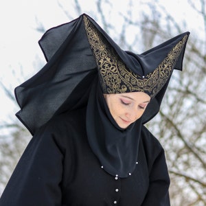 Puede incluir: Una mujer con una capa negra con capucha y un diseño estampado en oro y negro. La capucha es puntiaguda y tiene un velo largo y fluido.