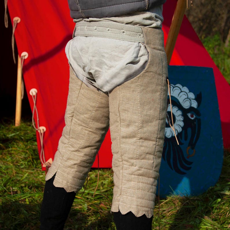 May include: A pair of beige quilted leg armor with scalloped edges. The armor is worn over a white undergarment and a brown belt. The armor is shown against a red background.