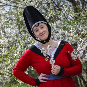 May include: A woman wearing a red dress with a black collar and a black hat with a silver trim. She is holding a bouquet of white flowers in her right hand. The background is a blurred image of white flowers.