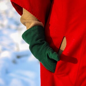 May include: A close-up of a person wearing a red coat with a green mitten. The mitten is covering a tan glove. The coat has a pocket. The background is a snowy, winter scene.