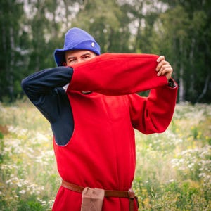 Op de afbeelding: Een felrode tuniek met lange mouwen en een bruine leren riem, gecombineerd met een donkerblauw onderkledingstuk met lange mouwen en een bijpassende blauwe vilten hoed met een klein goudkleurig embleem aan de zijkant als historisch kostuum.