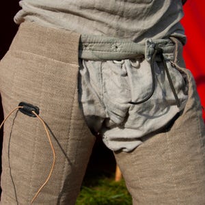 May include: A pair of brown linen pants with a black leather belt and a brown leather strap. The pants have a unique design with a flap that covers the front of the pants.