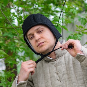 May include: A black quilted arming cap with vertical stitching and side flaps that tie under the chin. The cap features a light-coloured interior lining and is constructed from a durable fabric, designed to be worn securely around the head.
