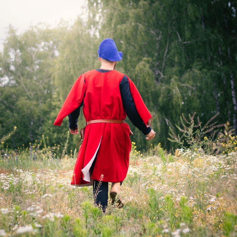 Op de afbeelding: Een felrode mouwloze tuniek gedragen over een donker shirt met lange mouwen, gecombineerd met een blauwe vilten muts en een bruine leren riem. Het kledingstuk heeft een lang, vloeiend ontwerp dat tot onder de knie&euml;n reikt.