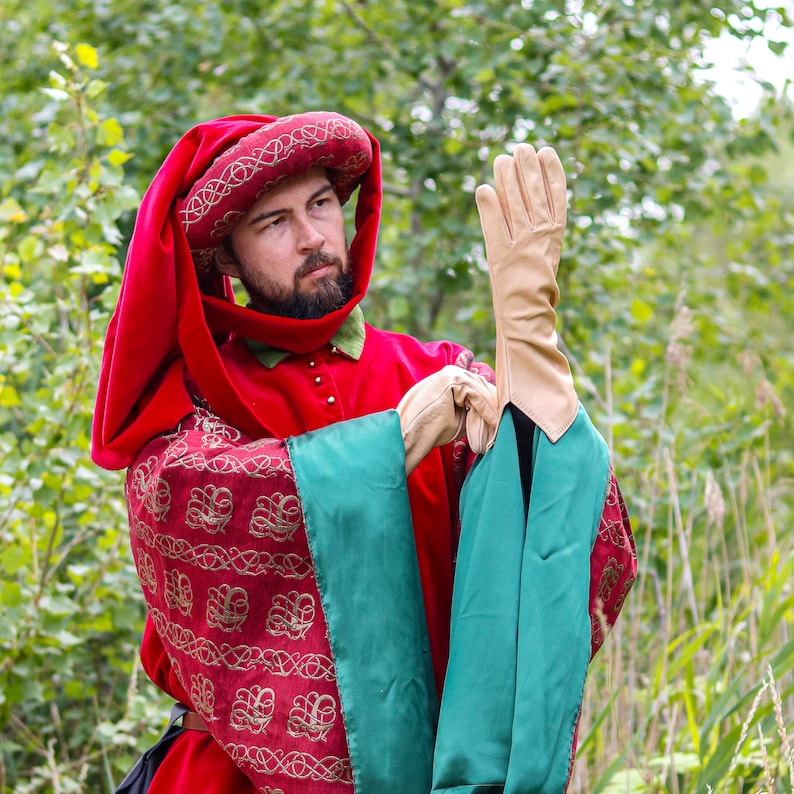 May include: A person wearing a red hooded robe with gold embroidery and green accents, holding a pair of brown leather gloves.