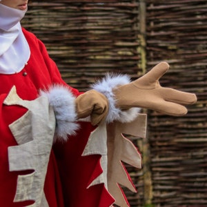 May include: A person wearing a red and white costume with fur-trimmed gloves. The gloves are brown and have a white fur cuff. The person is standing in front of a woven wall.