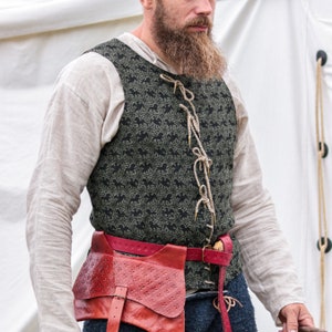 Könnte beinhalten: Ein Mann trägt eine grün-schwarze gemusterte Weste mit einem roten Ledergürtel und einer braunen Ledertasche. Die Weste hat einen Schnürverschluss mit einer braunen Schnur.