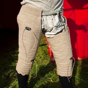 May include: A pair of beige linen leg armor with leather straps and buckles. The armor is quilted and has a textured surface. The armor is worn over pants and is secured with straps.