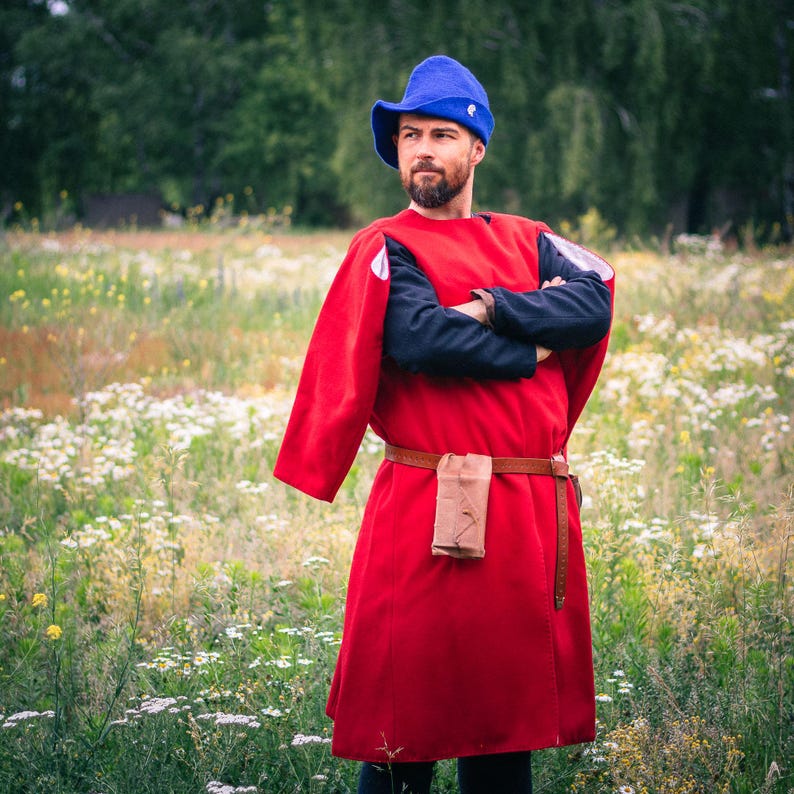 Op de afbeelding: Een felrode tuniek over een zwart shirt met lange mouwen, gecombineerd met een blauwe vilten hoed. Een bruine leren riem wordt rond de taille gedragen, met een klein bruin tasje aan de voorzijde bevestigd. Gedragen met een donkere broek.