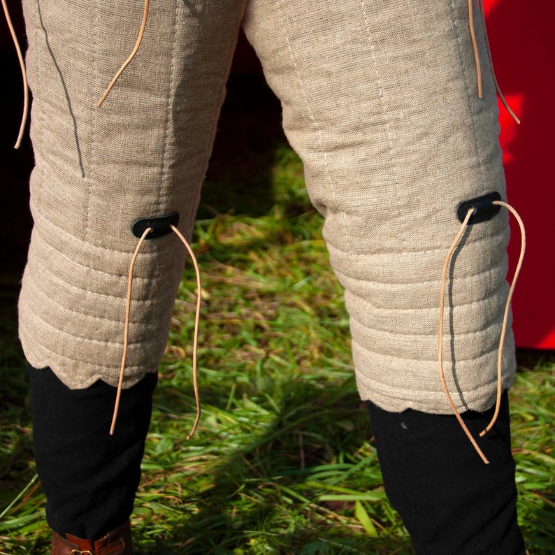May include: A pair of beige linen knee-length leggings with brown leather straps and buckles. The leggings are quilted and have a scalloped edge at the bottom.