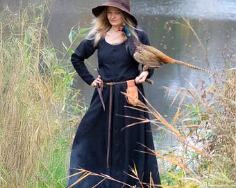 May include: A long-sleeved black floor-length dress paired with a brown pointed felt hat. A thin brown leather belt is worn at the waist, featuring a small tan leather pouch and a dark tool attached to the side. A pheasant is perched on one arm.