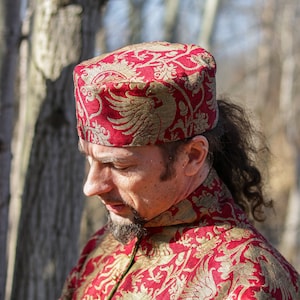 May include: A man wearing a red and gold patterned hat and matching long-sleeved shirt. The hat has a pointed top and a wide brim. The shirt has a high collar and a floral design.