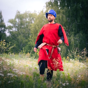 Op de afbeelding: Een felrode mouwloze tuniek gedragen over een zwart shirt met lange mouwen en een zwarte broek. De outfit bevat een blauwe vilten hoed, een bruine leren riem met een klein tasje en een bruine leren schede bevestigd aan de taille.
