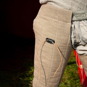 May include: A pair of tan linen pants with a black leather strap and a brown leather cord. The pants have a quilted design and are worn by a person.