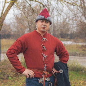 May include: A red medieval tunic with long sleeves and a high collar, fastened with green ties. The tunic is paired with blue pants, a brown belt, and a red and gray hat. The person is holding a black cloak and a sword.
