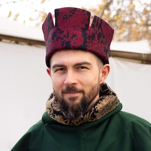 Peut inclure: Un chapeau rouge et noir à motifs avec un design de couronne unique. Le chapeau est porté avec un vêtement vert et un col à motifs noirs et dorés. L'image est un gros plan de la personne portant le chapeau.