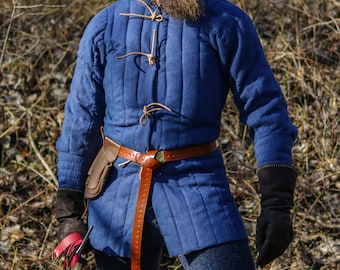 May include: A blue quilted gambeson featuring vertical stitching, a high collar, and light brown leather cord closures. A brown leather belt with a series of holes is worn around the waist, accompanied by a small leather pouch and dark gloves.