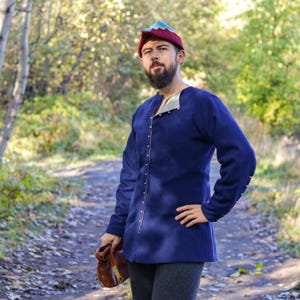May include: A man wearing a blue tunic with a brown leather pouch and a red and blue hat. The tunic has a high collar and is fastened with silver buttons. The man is standing in a forest setting.