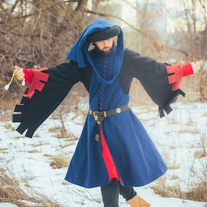 May include: A medieval costume in blue, black, and red, with a matching hood and a black belt with gold-coloured details. The costume is worn in a snowy outdoor setting. The tunic is knee-length, and the sleeves are long and flared.