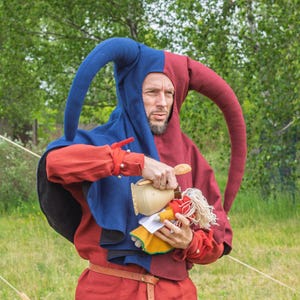 May include: A person wearing a red and blue jester costume with a long, curved hood. The costume includes a red tunic, a brown belt, and a wooden cup. The person is holding a doll with a red dress and blonde hair.