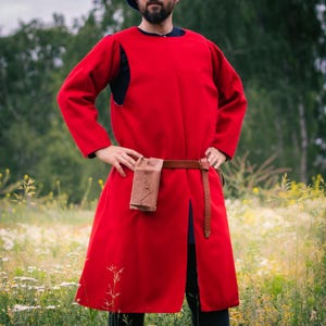 Op de afbeelding: Een rood tuniek met lange mouwen en knielengte, voorzien van een ronde halslijn en een centrale split aan de voorzijde. Het kledingstuk is in de taille ingesnoerd met een bruine leren riem waaraan een klein, rechthoekig bruin leren tasje hangt.