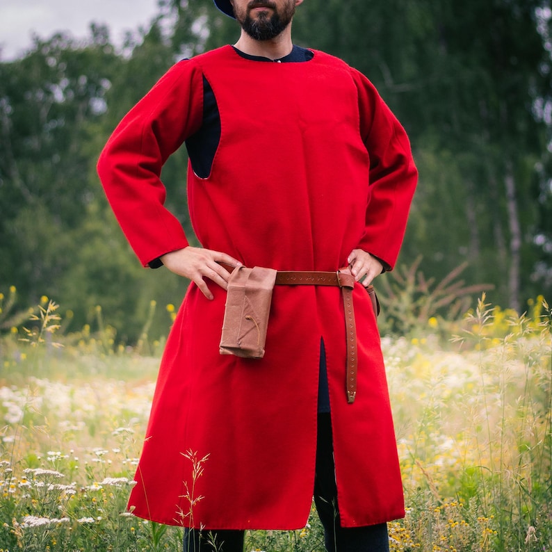 Op de afbeelding: Een rood tuniek met lange mouwen en knielengte, voorzien van een ronde halslijn en een centrale split aan de voorzijde. Het kledingstuk is in de taille ingesnoerd met een bruine leren riem waaraan een klein, rechthoekig bruin leren tasje hangt.