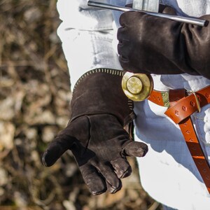 May include: A person wearing a white shirt and brown leather gloves. The gloves have a decorative stitching detail. The person is wearing a brown leather belt with a metal buckle and a gold-colored decorative piece.