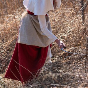 Puede incluir: Una mujer con un vestido blanco y rojo est&aacute; usando una hoz para cortar hierba seca en un campo.