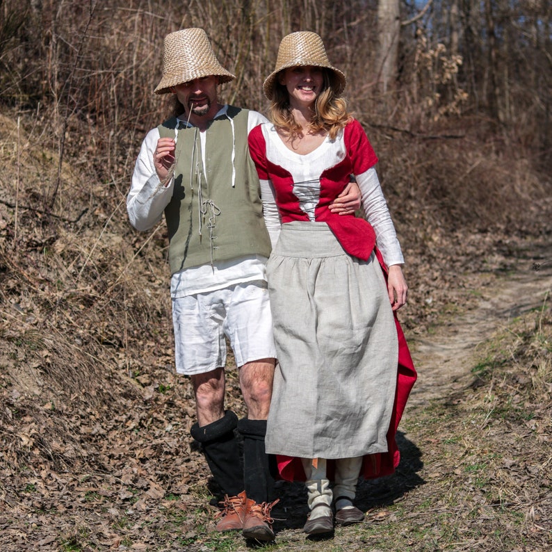 Puede incluir: Una pareja vestida con sombreros de paja y ropa de estilo medieval. La mujer lleva un vestido rojo y blanco con una falda larga. El hombre lleva un chaleco verde y pantalones blancos. Caminan por un sendero en un bosque.