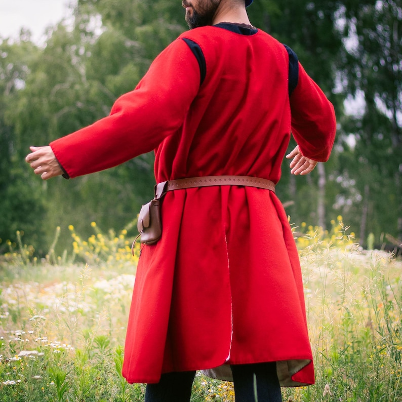 Op de afbeelding: Een rood tuniek met lange mouwen, een zwarte kraag en zwarte afwerking bij de schouders. Een bruine leren riem zit om de taille en houdt een klein bruin leren tasje aan de rechterkant vast. Het kledingstuk valt tot halverwege de bovenbenen.