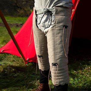 May include: A pair of beige linen leg armor with black straps and brown leather laces. The armor is worn over pants and has a quilted design.