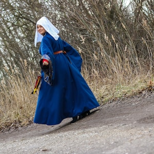 May include: A woman wearing a blue robe with a white head covering walks along a dirt path. The robe has a brown belt and a gold and red trim. The path is surrounded by tall, dry grass.