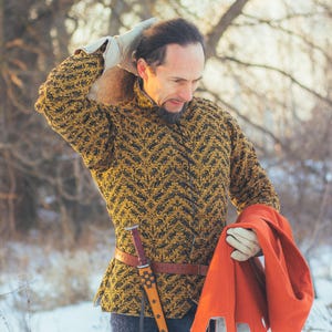 May include: A black and gold patterned medieval-style jacket with a high collar. The jacket is worn with a brown belt and a decorative dagger. The person is also wearing white gloves and holding an orange cloak. The jacket is likely made of quilted fabric.