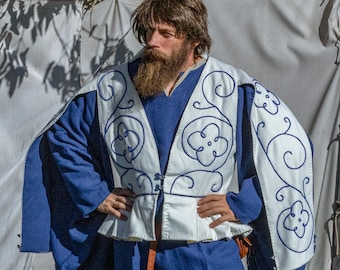 Puede incluir: Un hombre lleva una túnica azul con un chaleco blanco con un diseño floral. El chaleco tiene un ribete decorativo y una fila de botones en la parte delantera.